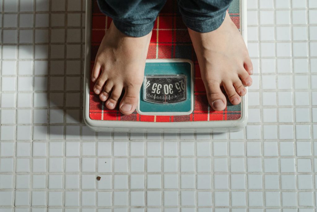 How Much Weight Can You Lose When Fasting? From above crop anonymous barefoot child in jeans standing on weigh scales on tiled floor of bathroom. How Much Weight Can You Lose When Fasting?