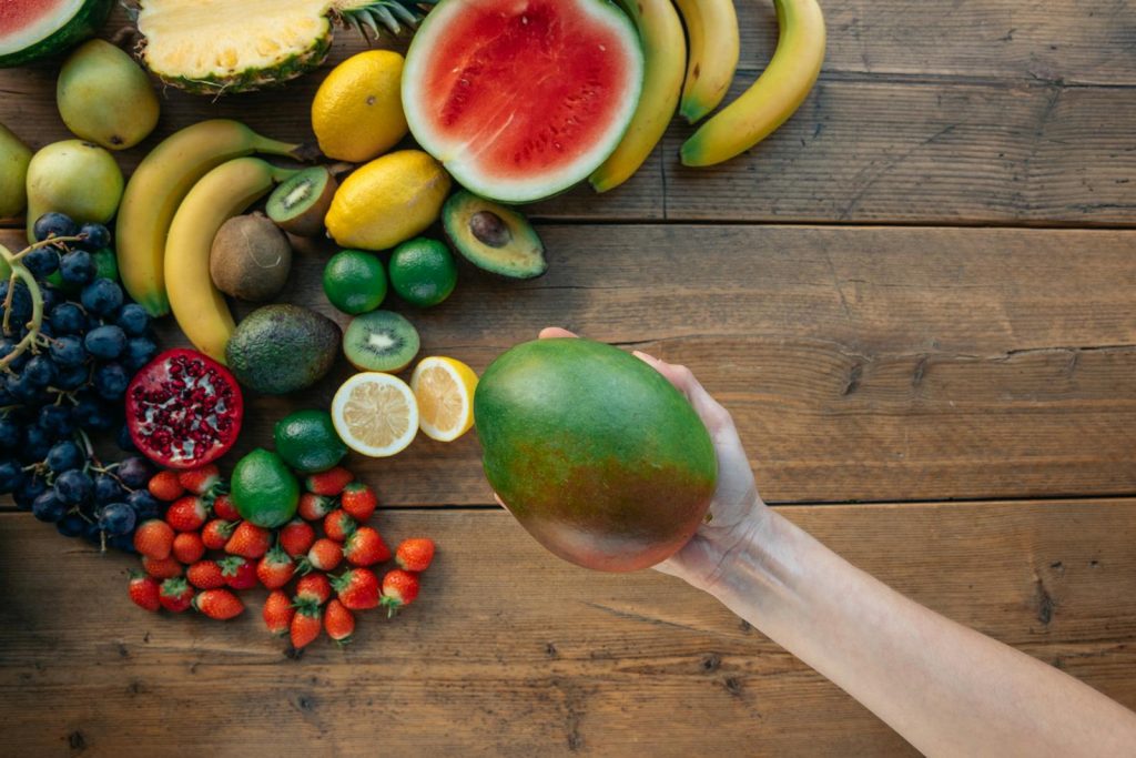 Diverse fresh fruits displayed on a wooden surface with a hand holding a mango.
